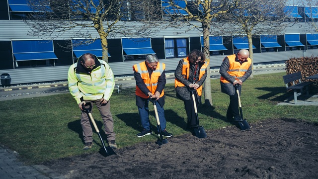 Four people in safety vests digging with shovels during a ceremonial groundbreaking in front of a building