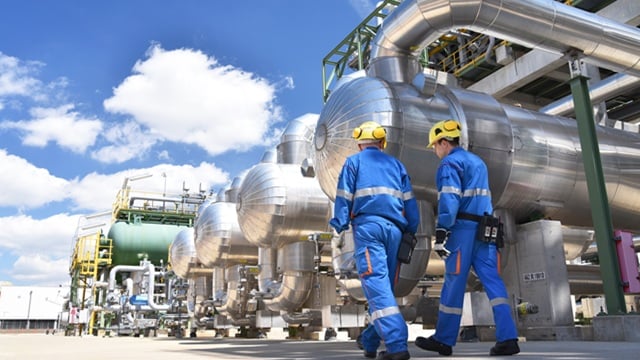 Two industrial workers in blue uniforms and yellow helmets inspect large metal pipes and machinery outdoors.